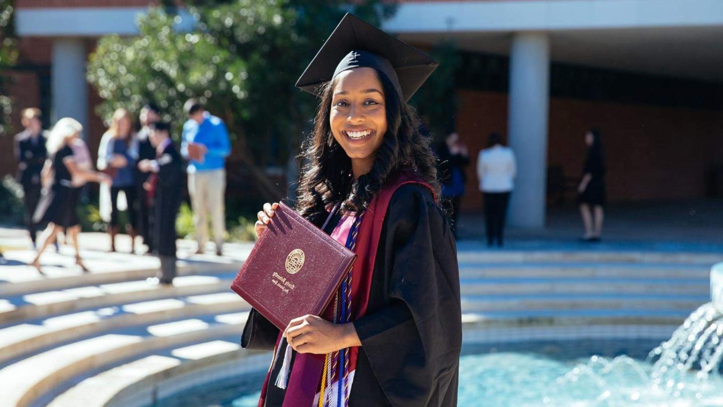 Rachel Daniel holding a degree in cap and gown, in front of Trinity's fountain